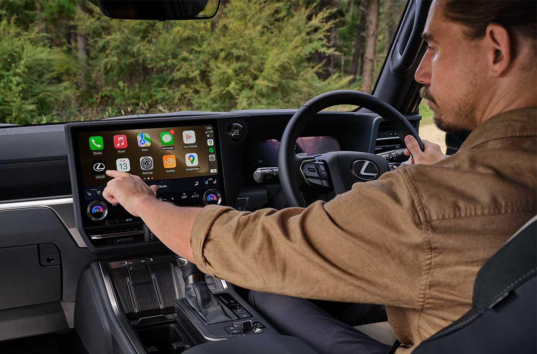 A passenger is shown sitting in the cockpit of the Lexus GX. He sits in the drivers seat, one hand holding the steering wheel, while the other touches the multi-media screen. The view from the front window shows greenery.