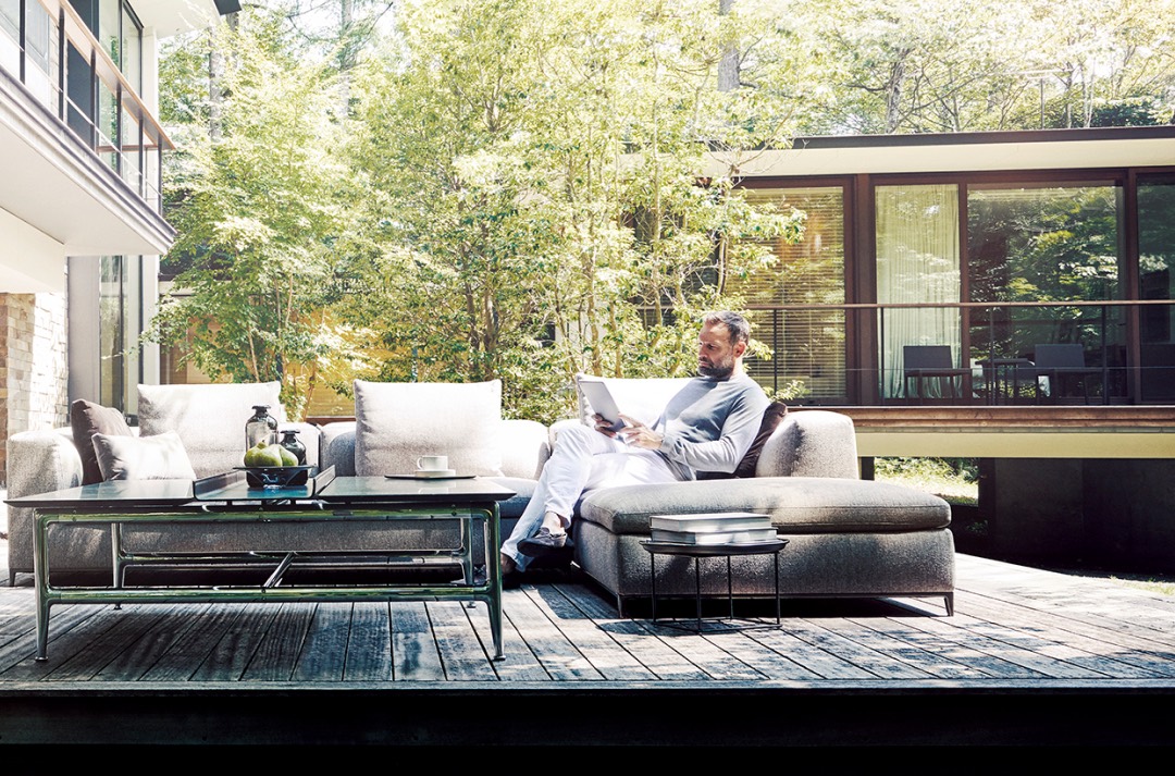 Man sits on an outdoor couch, reading a paper amongst green trees.