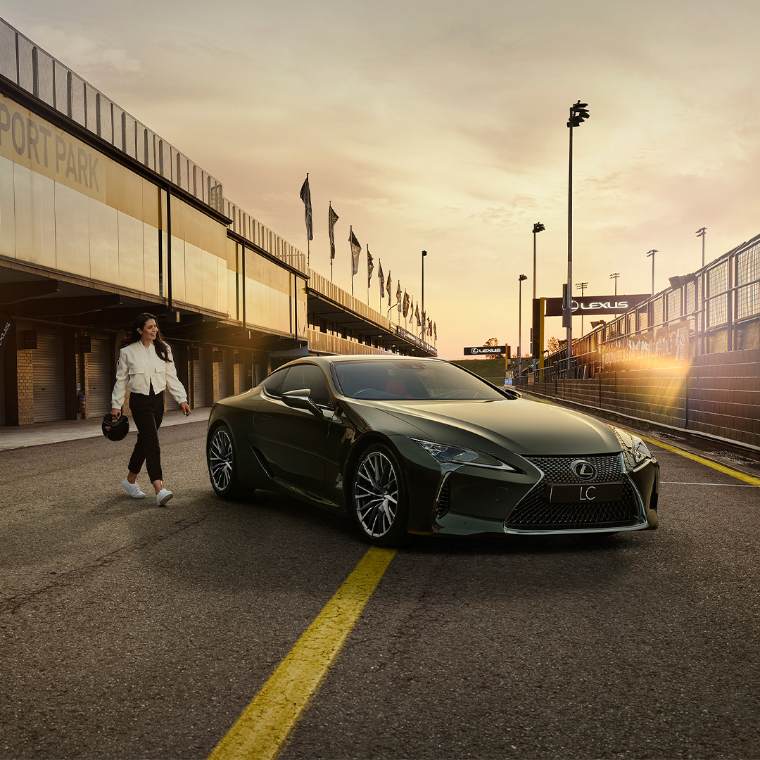 A woman dressed in a white jacket and black pants and holding a helmet walks towards an LC coupe parked in a pit lane.