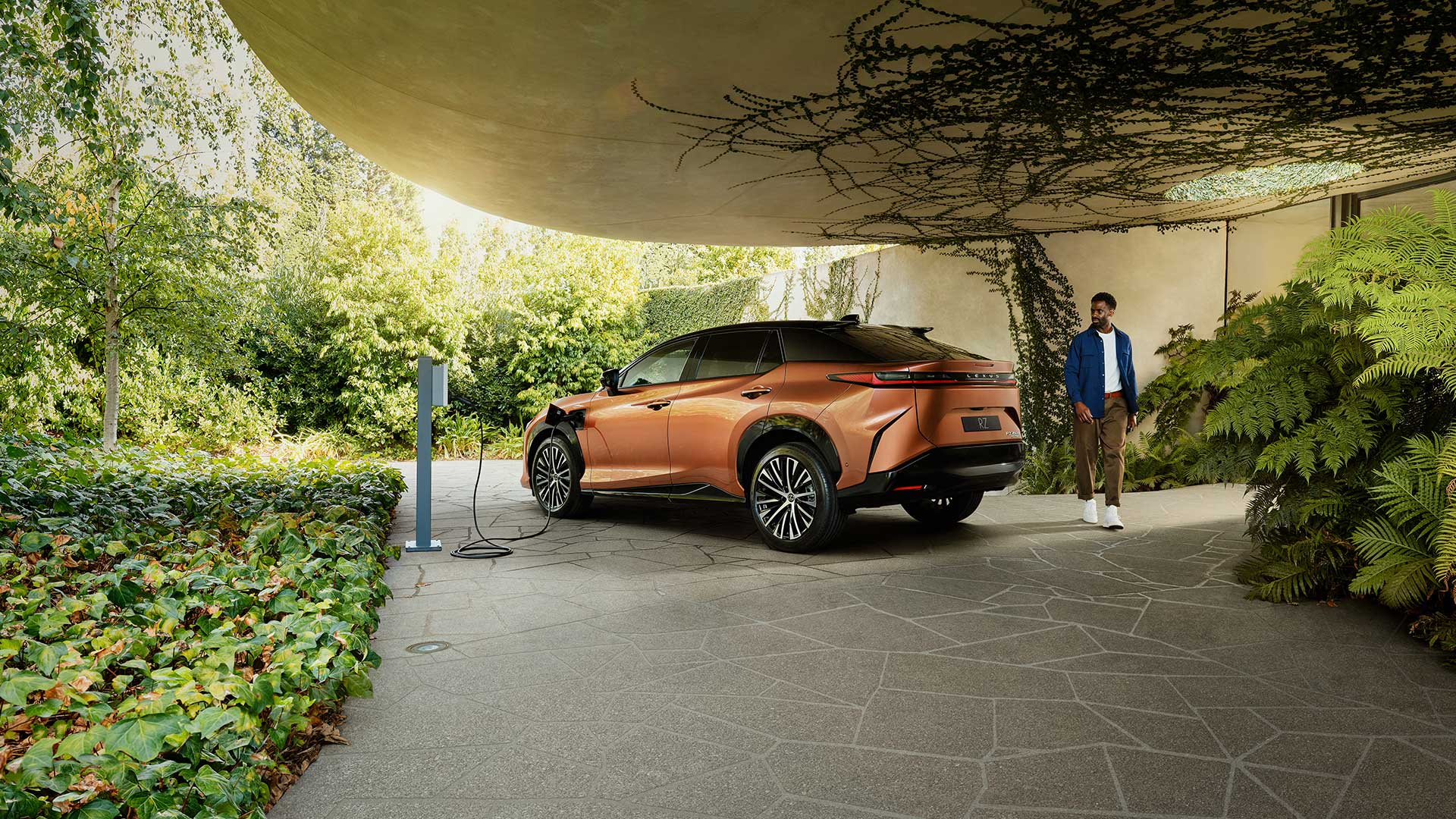 An copper Lexus RZ is parked and charging in a leafy green, vine-filled driveway. A man is seen walking around the rear of the car.